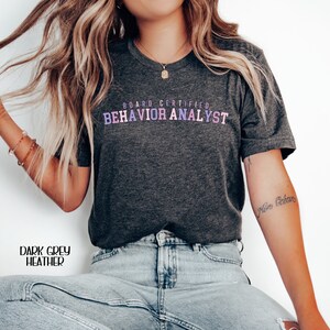 May include: A dark grey heather t-shirt with the text "BOARD CERTIFIED BEHAVIOR ANALYST" printed in rainbow colors.