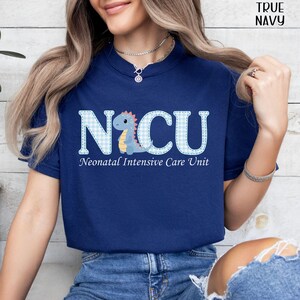 May include: A navy blue t-shirt with the text "NICU" in a light blue gingham pattern, featuring a cartoon dinosaur. Below the text is "Neonatal Intensive Care Unit" in a simple font. The words "True Navy" are in the top right corner.