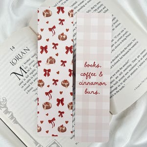 Könnte beinhalten: Zwei Lesezeichen liegen auf einem offenen Buch. Eines hat ein Muster aus roten Schleifen und Gebäck auf weißem Hintergrund. Das andere hat ein rosa Vichy-Muster mit den Worten "books, coffee & cinnamon buns."