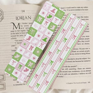 May include: Two decorative bookmarks. One has a repeating pattern of pink and green squares with whimsical icons. The other has a striped design with pink and green bows. Both are on top of an open book.