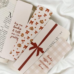 Könnte beinhalten: Eine Sammlung dekorativer Lesezeichen mit verschiedenen Designs. Ein Lesezeichen trägt den Text "buy me books & tell me I'm pretty". Ein anderes zeigt ein rotes Schleifendesign. Andere haben Zimtschnecken- und Schleifenmuster. Ein Lesezeichen sagt "books, coffee & cinnamon buns."