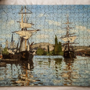 May include: A vintage jigsaw puzzle depicting two ships sailing on a calm blue body of water. The ships are brown with white sails and are reflected in the water. The background features a cityscape with buildings and trees. The puzzle is complete and has a light wood frame.