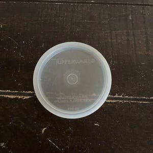 May include: A clear, round Tupperware lid with the brand name and "Made in U.S.A." printed on it. The lid is translucent and has a slightly raised edge. The text "Tupperware Orlando, Florida, USA" is also visible.