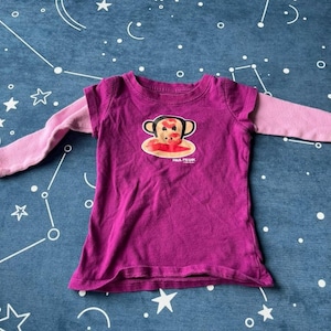 Może przedstawiać: Magenta koszulka z długim rękawem z grafiką małpy z przodu. Rękawy są w kolorze jasnoróżowym. Pod grafiką małpy znajduje się napis "PAUL FRANK". Odzież dziecięca.