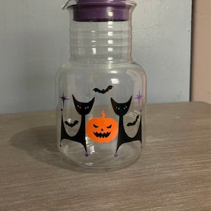 May include: A clear glass pitcher with a purple lid and a Halloween design featuring two black cats, a pumpkin, and bats.