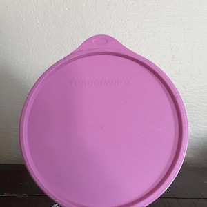 Puede incluir: Una tapa redonda de Tupperware de color lila claro. La tapa tiene un borde elevado y una pequeña pestaña. La palabra "Tupperware" está grabada en el centro. La tapa está hecha de plástico resistente y está diseñada para un recipiente.