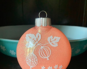 May include: A peach-colored Christmas ornament with a white glitter finish. The ornament features a white silhouette of a gooseberry plant with leaves and berries.
