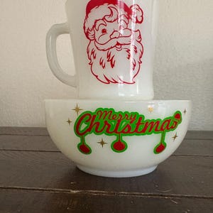 Może przedstawiać: Biały ceramiczny kubek z czerwonym motywem Świętego Mikołaja i biała miska z napisem "Merry Christmas" w kolorze zielonym i czerwonym. Miska ma czerwone wzory ozdób i złote gwiazdki.