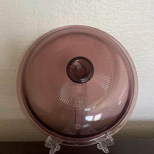 Puede incluir: Una tapa redonda de vidrio púrpura transparente con un pomo central. La tapa presenta un diseño radial que emana del pomo. La tapa se muestra en un soporte de plástico transparente, mostrando su diseño y color.