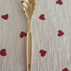 May include: A beige plastic spaghetti server with a slotted head and a long handle. The server is resting on a white cloth with a pattern of red hearts.