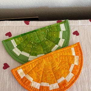 May include: Two handmade, quilted citrus slice-shaped trivets. One is green, the other orange, with white and patterned accents. The trivets are designed to resemble orange and lime slices, perfect for kitchen decor.