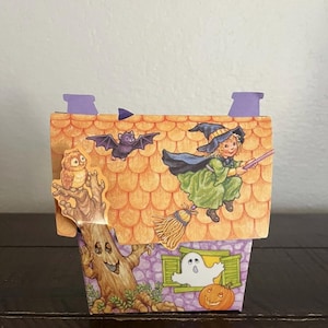 May include: A Halloween-themed paper house with a witch riding a broom, a bat, an owl, a ghost, and a jack-o'-lantern. The roof is orange with a scale pattern, and the house has purple accents. The overall design is whimsical and festive.