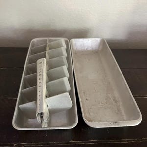 May include: Vintage metal ice cube tray with a rectangular shape and a divider. The tray is divided into individual ice cube compartments. The word "BUCK KUBE" is printed on the handle. The tray is made of metal and has a worn appearance.