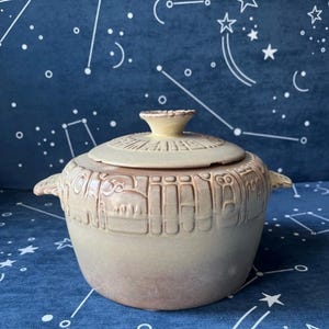 May include: A beige ceramic casserole dish with a lid. The dish has a textured design around the sides and a small handle on the lid. The dish has two handles on the sides.