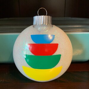 May include: A white glitter Christmas ornament with a stack of colorful semi-circles in blue, red, green, and yellow.