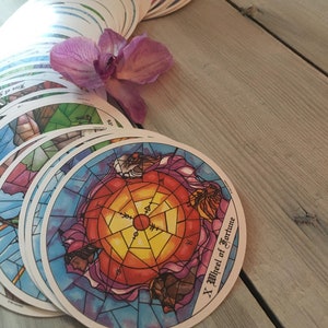 Puede incluir: Un mazo de cartas de tarot redondas con ilustraciones coloridas, incluida la carta "X Wheel of Fortune", dispuestas sobre una superficie de madera. Una flor de orquídea púrpura descansa cerca de las cartas, agregando un toque de belleza natural.