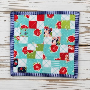 May include: A square quilted potholder featuring a patchwork design of various fabric squares in shades of turquoise, red, white, and lime green. The pattern includes floral motifs, cherries, and plaid prints, all finished with a dark blue polka dot border.