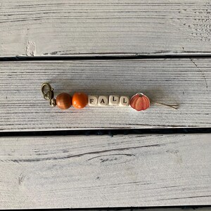 Op de afbeelding: Een sleutelhanger met een bronskleurige metalen sluiting, een gladde bruine houten kraal, een feloranje kraal, vier vierkante houten kralen met de zwarte letters FALL en een platte houten bedel in de vorm van een pompoen met oranje strepen.