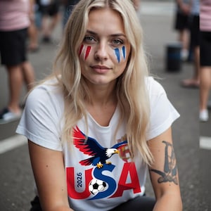 以下が含まれることがあります： 金髪で赤、白、青のフェイスペイントをした女性。白いTシャツを着ており、ワシ、サッカーボール、そして「USA 2026」の文字が描かれています。画像は屋外で撮影されました。