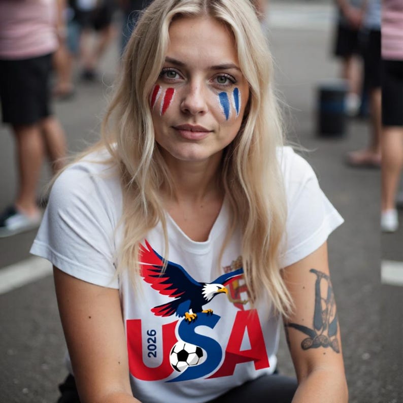 以下が含まれることがあります： 金髪で赤、白、青のフェイスペイントをした女性。白いTシャツを着ており、ワシ、サッカーボール、そして「USA 2026」の文字が描かれています。画像は屋外で撮影されました。
