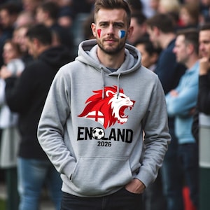 以下が含まれることがあります： 赤と白のライオンのデザインと、サッカーボールが付いた黒い文字で「ENGLAND」と書かれたグレーのフード付きスウェットシャツ。 年号「2026」が下に印刷されています。 画像の人物は顔にペイントを施しています。
