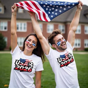 May include: Two people wearing white t-shirts with a USA flag and soccer ball graphic, and the text "USA 2026". They are holding up an American flag, with face paint in red, white, and blue. The background is a blurred outdoor scene.