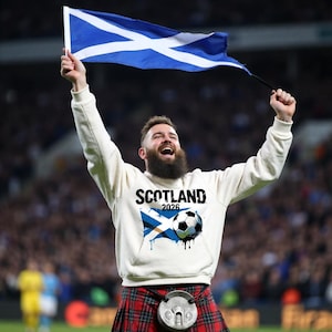 May include: A person wearing a white hoodie with "SCOTLAND 2026" printed on it, holding a Scottish flag. The hoodie features a Scottish flag design with a soccer ball. The person is also wearing a traditional Scottish kilt.