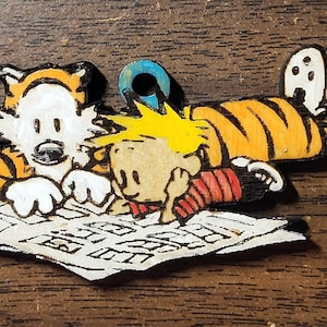 May include: A wooden ornament featuring the cartoon characters Calvin and Hobbes. Calvin is lying on his stomach, reading a newspaper with Hobbes, the tiger, beside him. The ornament is painted in black, white, orange, and yellow.