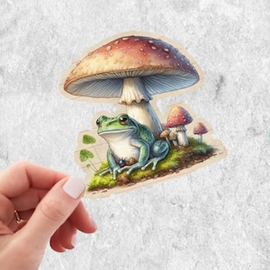 May include: A sticker featuring a whimsical illustration of a green frog sitting on moss, beneath a large mushroom with a red and white spotted cap. Small mushrooms are also present. The sticker has a vintage, hand-drawn aesthetic.