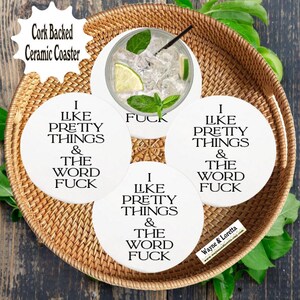 May include: A set of four white ceramic coasters with cork backing, each featuring the text "I LIKE PRETTY THINGS & THE WORD FUCK" in black. The coasters are arranged on a woven tray, with a drink on one coaster.