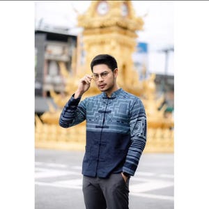 May include: A blue and navy button-up shirt with a geometric pattern. The shirt has a mandarin collar and long sleeves. The sleeves have a striped design. The shirt is paired with gray pants.