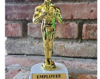 May include: A gold-colored trophy featuring a male figure in a standing pose, mounted on a white base with a gold plaque. The plaque reads "EMPLOYEE OF THE MONTH" in black text. The background is a brick wall.