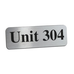 May include: A rectangular silver metal sign with rounded corners. The sign has the words "Unit 304" in bold black font. The sign is set against a white background.