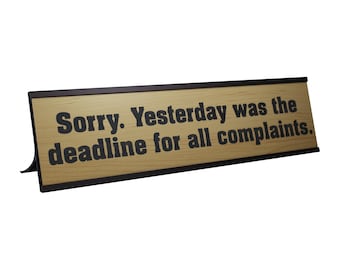 May include: A wooden desk sign with the text "Sorry. Yesterday was the deadline for all complaints." The sign has a black frame and a light brown wood grain background. The text is in a bold, black font.