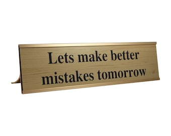 May include: A rectangular desk sign with a gold frame and a light wood grain appearance. The sign reads "Lets make better mistakes tomorrow" in black text. The sign has a small stand on the back.