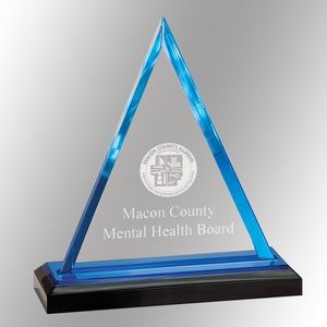 May include: A clear acrylic triangle award with blue edges and a black base. The text "Macon County Mental Health Board" is printed on the front, along with a circular seal. The award is designed to stand upright.