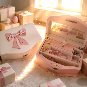 May include: A light pink jewelry box with a hinged lid featuring a large pink bow and the name Amelia in white script. The open box reveals a mirror, a rotating ballerina figurine, and multiple compartments containing various jewelry pieces.