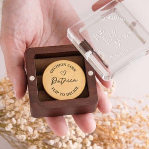 May include: A round gold-colored metal decision coin with the text "DECISION COIN", a heart symbol, "Patrica", and "FLIP TO DECIDE" engraved on it, housed in a square dark brown wooden box with a clear lid featuring "THE WILLIAMS", "Who Will Decide?", and "JOSEPH & PATRICIA" etched on the surface.