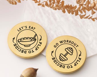 May include: Two circular gold-toned metal decision coins. One coin features a bowl of food with the text "LET'S EAT" and "FLIP TO DECIDE". The other coin displays a dumbbell icon with the text "GO WORKOUT" and "FLIP TO DECIDE".