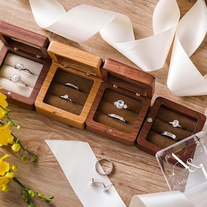 May include: Four square wooden ring boxes with hinged lids, each containing two silver rings. The boxes feature velvet inserts in brown or cream. A clear acrylic lid with the engraved text A & B 2025 is placed nearby, alongside loose silver rings and a pair of earrings.