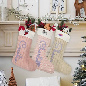 May include: Three knitted Christmas stockings hanging in a row. The left is pink with the name Cecilia, the middle is light pink with Charlotte, and the right is beige with Shanna. Each has a white cuff with embroidered icons and small plush toys.