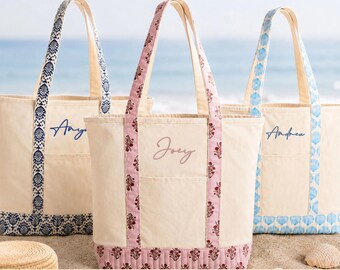 May include: Three off-white canvas tote bags with exterior pockets and patterned straps. The left bag has blue trim and the name Anya, the center bag has pink floral trim and the name Joey, and the right bag has light blue trim and the name Andrea.