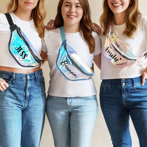 May include: Three iridescent fanny packs with black zippers and adjustable straps. Each bag features a custom name printed in black script: Jess, Brianna, and Emily. The bags have a reflective, holographic finish that shifts colors under light.