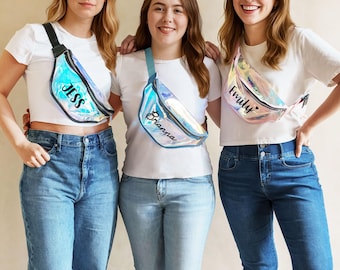 May include: Three iridescent fanny packs with black zippers and adjustable straps. Each bag features a custom name printed in black script: Jess, Brianna, and Emily. The bags have a reflective, holographic finish that shifts colors under light.