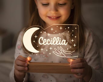 May include: A custom acrylic night light featuring a warm glowing crescent moon, a hand reaching toward stars, and the name Cecillia engraved in white script. The clear acrylic panel is mounted on a rectangular light-colored wooden base.