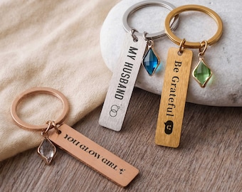 May include: Three metal keychains with rectangular tags. One silver tag reads MY HUSBAND with a ring icon and a blue crystal charm. One gold tag reads Be Grateful with a smiley face and green crystal. One rose gold tag reads YOU GLOW GIRL.