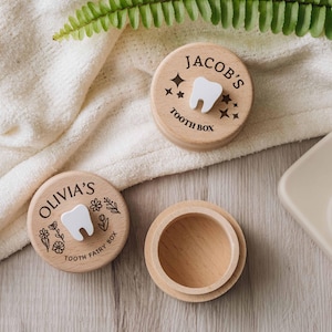 May include: Two round wooden boxes with lids and one open wooden base. One lid features a white tooth icon, stars, and the text "JACOB'S TOOTH BOX". The other lid features a white tooth icon, floral designs, and the text "OLIVIA'S TOOTH FAIRY BOX".