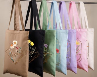 May include: A row of seven canvas tote bags in tan, black, sage green, light blue, lavender, pink, and cream. Each bag features a gold-outlined diamond frame containing a floral illustration and the printed text Lilian and BRIDESMAID.