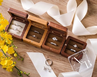 May include: Four square wooden ring boxes in varying shades of brown, each containing two silver-toned rings. A clear acrylic lid with the text A & B 2025 is placed nearby. The arrangement includes yellow flowers and white ribbon on a wood surface.