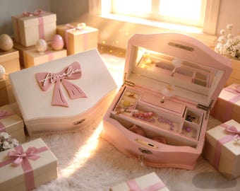 May include: A light pink jewelry box with a ballerina figurine inside, featuring a mirror, multiple compartments, and a pink bow on the lid with the name Amelia written in white script. The box is surrounded by several gift boxes with pink ribbons.